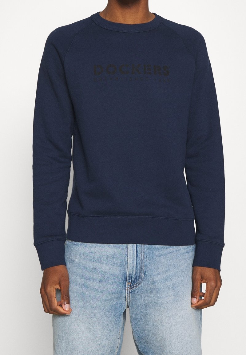 Námořnická modrá mikina s raglánovými rukávy, měkká textura, kulatý límec a černé reliéfní logo "DOCKERS" na přední straně. Diskrétní ribované manžety a lem.