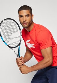 Rød bomull T-skjorte med logo av en hvit krokodille, kombinert med et svart og blått tennisracket, som viser en atletisk kroppsbygning.