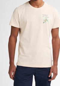 Camiseta de algodón beige con mangas cortas, que presenta un pequeño estampado botánico colorido y un logo en el área del bolsillo del pecho.