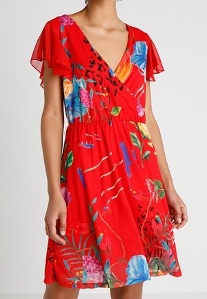 Vrouw in een rode jurk tot op de knie met een kleurrijke bloemenprint en wijd uitlopende mouwen, staand met haar armen ontspannen langs haar zij.