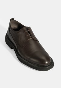 Chaussure habillée en cuir brun avec une finition lisse, bout rond et fermeture à lacets. Dispose d'une semelle en caoutchouc noir texturée et de détails de couture subtils.