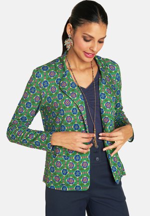 Grüner Blazer mit blau-rosafarbenem Blumenmuster. Taillierte Passform, mit Revers und zwei vorderen Taschen. Über einem navyfarbenen Oberteil getragen.