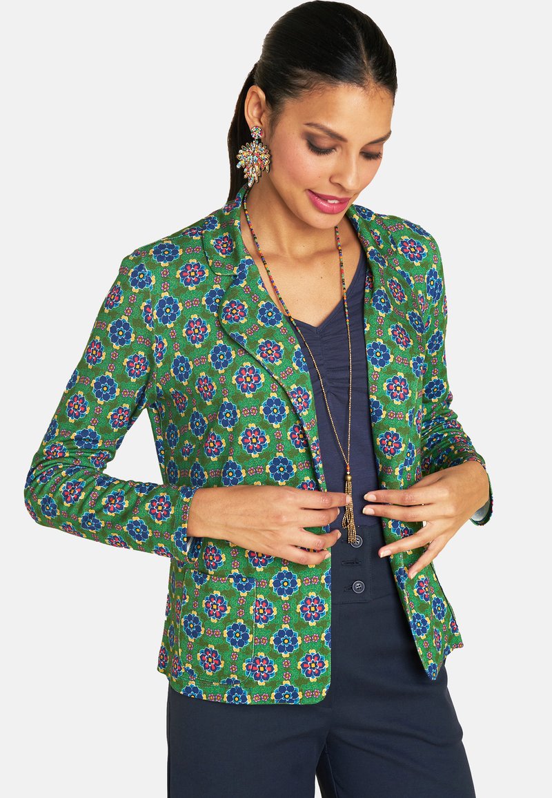 Green patterned blazer with blue and pink floral designs. Tailored fit, notched lapels, and two front pockets. Worn over a navy top.