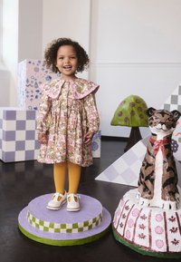 Dieťa v kvetinových šatách s veľkým ružovým golierom, žltými pančuchami a bielymi topánkami, stojace na fialovom podstavci so zelenými prvkami.