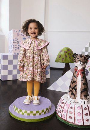 Laps lillelillelise kleidiga, millel on suur roosa krae, kollased sukkpüksid ja valged kingad, seisab lilla pjedestaali peal rohelist värvi detailidega.