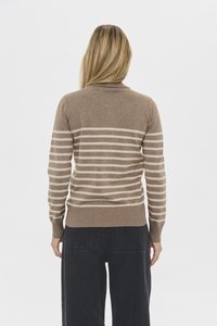 Kobieta o długich blond włosach, ubrana w brązowo-beżowy sweter w paski i czarne spodnie, stojąca tyłem na jednolitym tle.