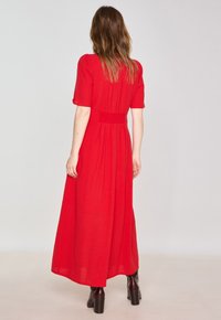 Red maxi dress with short sleeves, textured fabric, fitted waist, pleated back detail, and a flowing silhouette, paired with brown heeled boots.