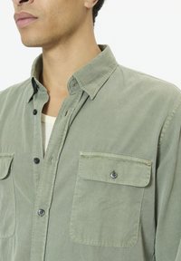 Mees, kes kannab helerohelist nööpidega särki, millel on kaks esimest taskut, krae on lahti ja valge aluspesu paistab kaeluses.