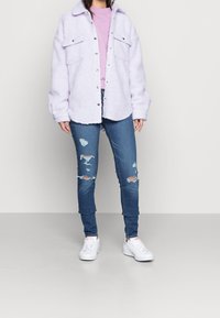Persona vestida con una chaqueta de sherpa lila sobre una blusa rosa clara, jeans ajustados azules desgastados y zapatillas blancas, de pie sobre un fondo plano.