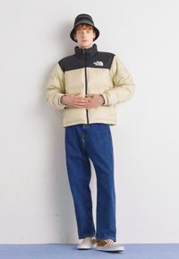 Pufferjack met een beige ondergedeelte en een zwart bovengedeelte, gedragen over een blauwe denim jeans. Accessoires zijn een zwarte bucket hat en geblokte schoenen.