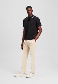 Černé polo tričko s bílým límečkem a vyšitými detaily, spárované se světle béžovými kalhotami a bílými teniskami.