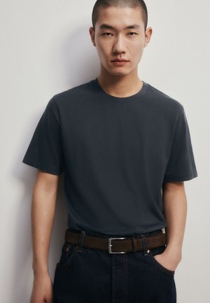 Jonge man met kort haar, draagt een donkergrijs T-shirt dat in een donkere spijkerbroek met een bruine riem is gestopt, staand tegen een effen witte muur.