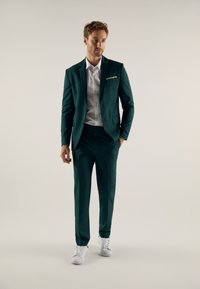 Grüner Anzug mit taillierter Passform, einreiher Jacke, Revers, und einer Einstecktuch. Kombiniert mit einem weißen Hemd und weißen Sneakers.