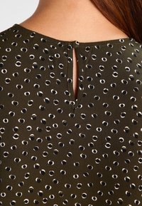 Tessuto verde scuro con una fantasia a pois caratterizzata da cerchi neri e bianchi. Apertura a chiave sul retro con una chiusura a singolo bottone.