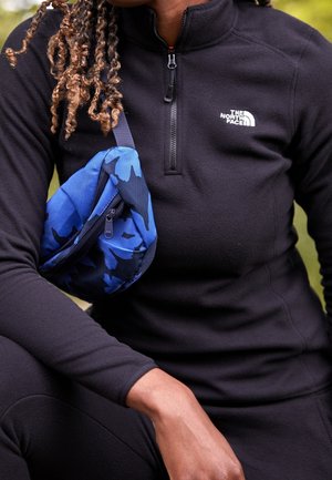 Persoon die een zwarte North Face-jas met rits draagt en buiten een taillezak met blauw camouflagepatroon over de borst houdt.