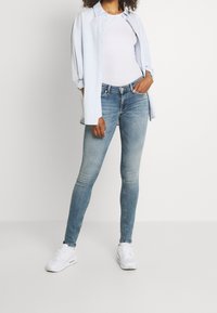 Jeans ajustados de mezclilla de color azul claro, con un acabado desgastado. Combinados con una camiseta blanca y una camisa abotonada azul claro. Zapatillas blancas.