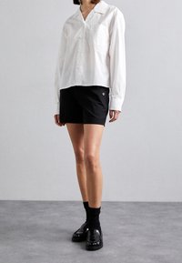Camicia bianca a maniche lunghe, morbida e con colletto, abbinata a pantaloni corti neri su misura con dettagli a bottoni. Mocassini neri e calzini.