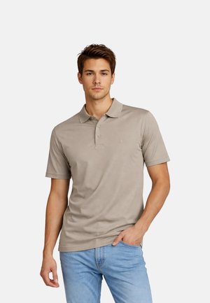 Junger Mann, der ein taupefarbenes kurzärmeliges Poloshirt und hellblaue Jeans trägt, steht mit einer Hand in der Tasche vor einem einfarbigen Hintergrund.