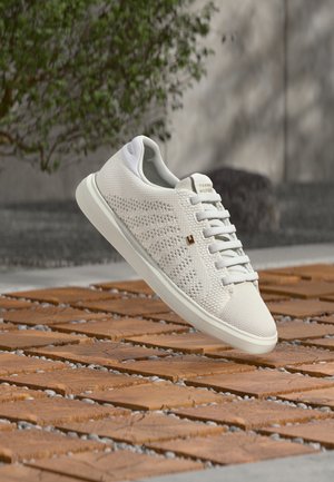 Fehér kötött sportcipő fűzővel és oldalon lévő logóval, lebegve egy barna csempézett kültéri felület felett, háttérben elmosódott zöld növényzettel és fallal.