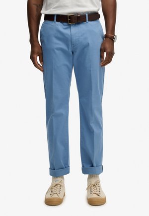 Homme portant un pantalon bleu clair avec revers, ceinture marron, chaussettes blanches et baskets en toile beige, debout avec les bras détendus le long du corps.