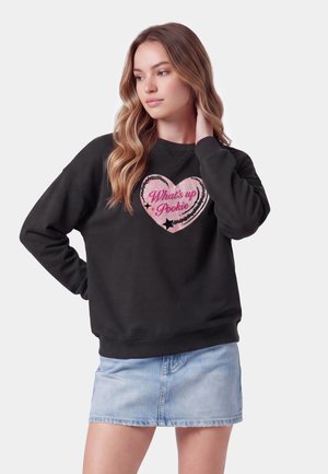 Giovane donna con capelli lunghi e ondulati che indossa una felpa nera con un cuore rosa e il testo "What's up Pookie" e una gonna di jeans azzurro chiaro.