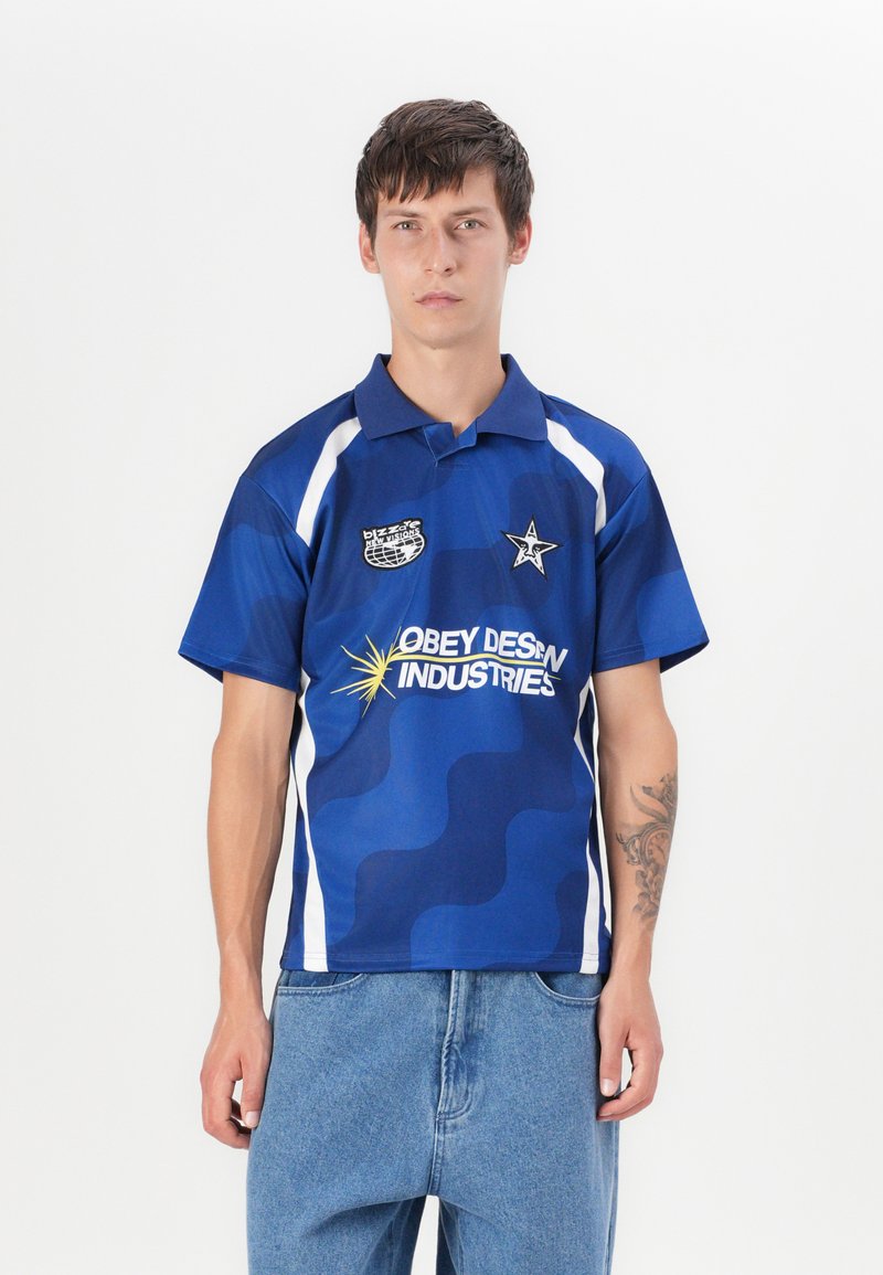 Blue patterned polo shirt with white accents, featuring a collar, logos, and a relaxed fit. Worn with light blue jeans.