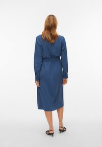 Femme aux cheveux mi-longs portant une robe bleue ceinturée, à manches longues et longueur genoux, ainsi que des sandales à talons noires, debout, de dos.