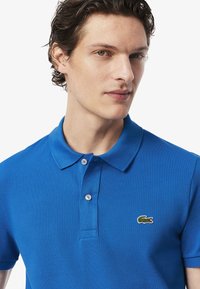 Polo azul hecho de tela texturizada, con cuello clásico, tapeta de dos botones y un pequeño logo de cocodrilo verde en el pecho.