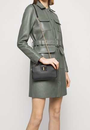 Žena v zelených kožených šatech s páskem drží malou černou koženou kabelku přes rameno se zlatým řetízkovým popruhem a sponou.