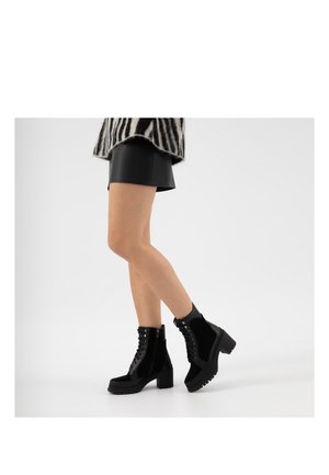 Femme portant des bottines noires à lacets avec talons épais, une mini-jupe noire et un pull à motif zèbre sur un fond blanc.