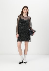Rochie neagră cu paiete, cu mâneci lungi transparente, până la genunchi, dintr-un material texturat, asociată cu o geantă plic neagră și șosete albe.
