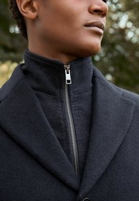 Cappotto nero con colletto alto e dettagli con zip, caratterizzato da un tessuto testurizzato e accenti lisci, che mette in risalto un design su misura.