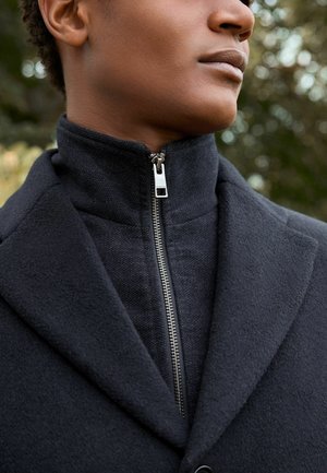 Abrigo negro con cuello alto y detalles de cremallera, que presenta una tela texturada y acentos suaves, mostrando un diseño ajustado.