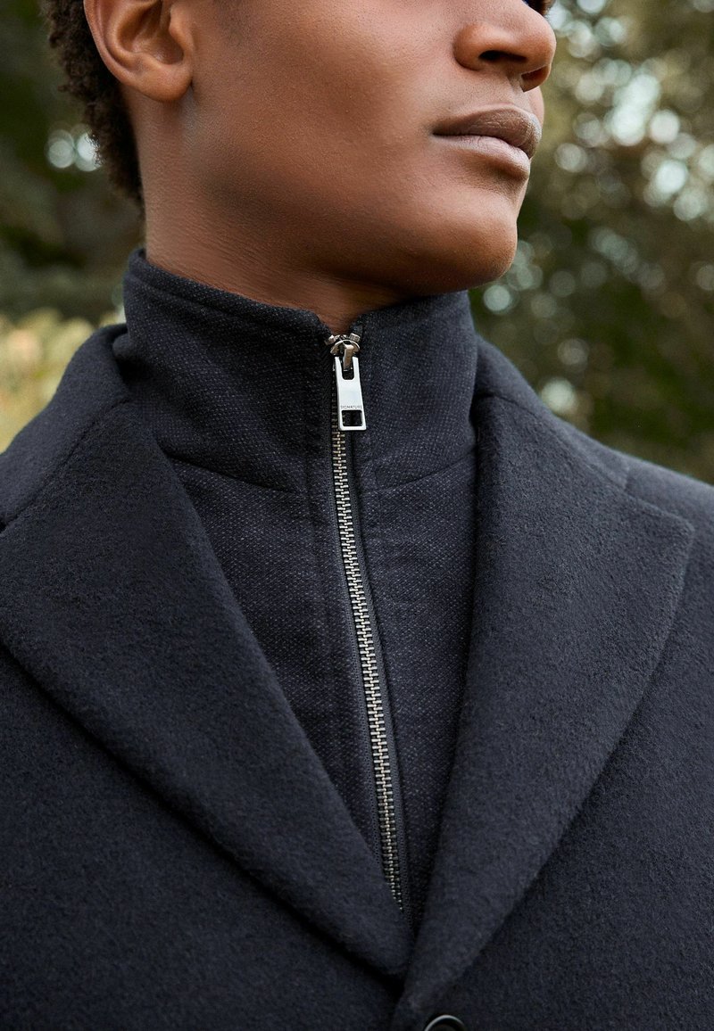 Cappotto nero con colletto alto e dettagli con zip, caratterizzato da un tessuto testurizzato e accenti lisci, che mette in risalto un design su misura.