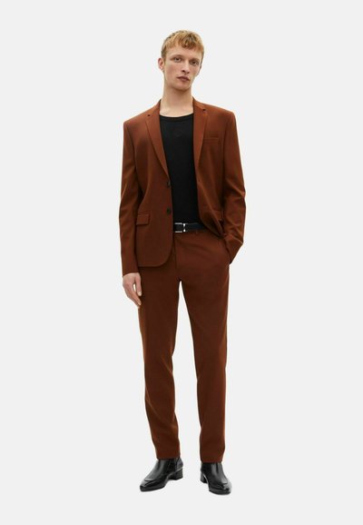 Brąski garnitur dopasowany w fasonie slim fit. Żakiet ma pojedynczy guzik i klapy w kształcie notcha, zestawiony z czarną bluzką i czarnymi butami.