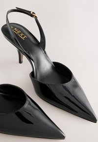 Escarpins slingback en cuir verni noir avec bouts pointus et talons aiguilles fins de hauteur moyenne sur une surface claire.