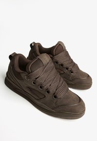 Par temno rjavih superg z debelimi vezalkami, mrežastimi paneli ter oblazinjenim jezikom in ovratnikom na beli podlagi.