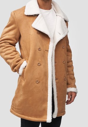 Man draagt een beige dubbelgeknoopt jas met witte schapenvachtkraag en manchetten, handen gedeeltelijk in de zakken, over een wit overhemd en zwarte broek.