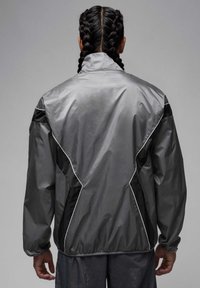Grå vindjacka i lätt material med kontrasterande svarta detaljer och vit piping längs sömmarna. Klassisk krage och avslappnad passform.