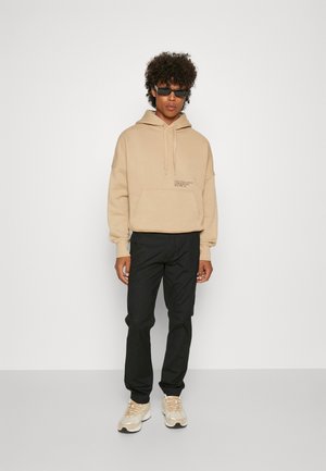 Felpa beige con cappuccio e cordino, abbinata a pantaloni neri e sneakers beige. Gli accessori includono occhiali da sole, creando un look casual.