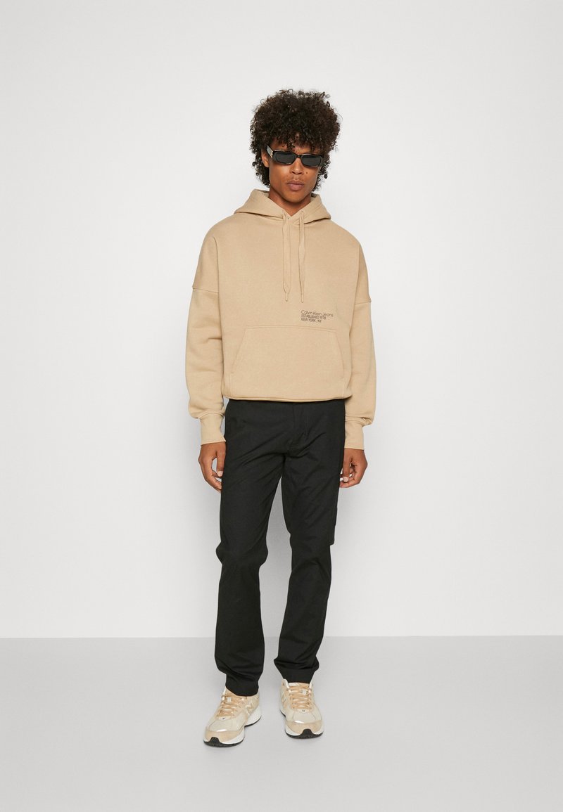 Felpa beige con cappuccio e cordino, abbinata a pantaloni neri e sneakers beige. Gli accessori includono occhiali da sole, creando un look casual.
