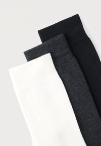 Τρία ζευγάρια διπλωμένων καλτσών σε λευκό, σκούρο γκρι και μαύρο, τοποθετημένα το ένα δίπλα στο άλλο σε ανοιχτό φόντο.