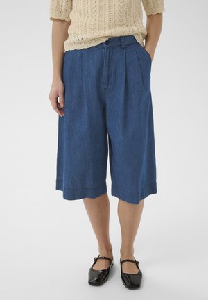 Frau trägt blaue, weit geschnittene, knielange Jeansshorts, beiges gestricktes Kurzarmtop und schwarze Mary-Jane-Schuhe.
