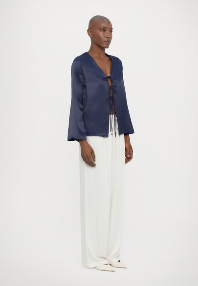 Blusa de satén azul marino con escote en V y detalle de lazada en la parte delantera, combinada con pantalones de pierna ancha blancos. Se coloca por debajo de las caderas y cuenta con mangas largas.