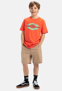 Camiseta de manga corta naranja con logo gráfico azul, pantalones cortos beige y zapatillas negras con detalles blancos. Combinado con calcetines negros.