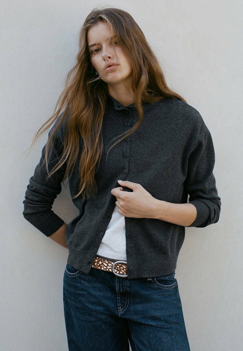 Jeune femme aux longs cheveux bruns portant un cardigan gris foncé boutonné, une chemise blanche, un jean bleu et une ceinture à motifs, se tient devant un fond uni.