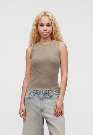 Jeune femme aux longs cheveux blonds bouclés porte un haut beige sans manches et un jean ample bleu clair, debout devant un fond clair uni.