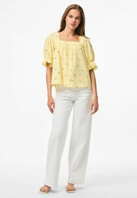 Blusa gialla con scollatura quadrata e maniche a palloncino corte, con piccoli motivi floreali. Abbinata a pantaloni bianchi a gamba larga e sandali.
