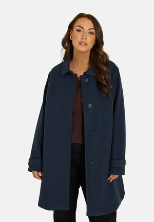 Donna con i capelli castani lunghi che indossa un cappotto blu scuro abbottonato sopra una maglia scura e pantaloni neri su uno sfondo semplice.