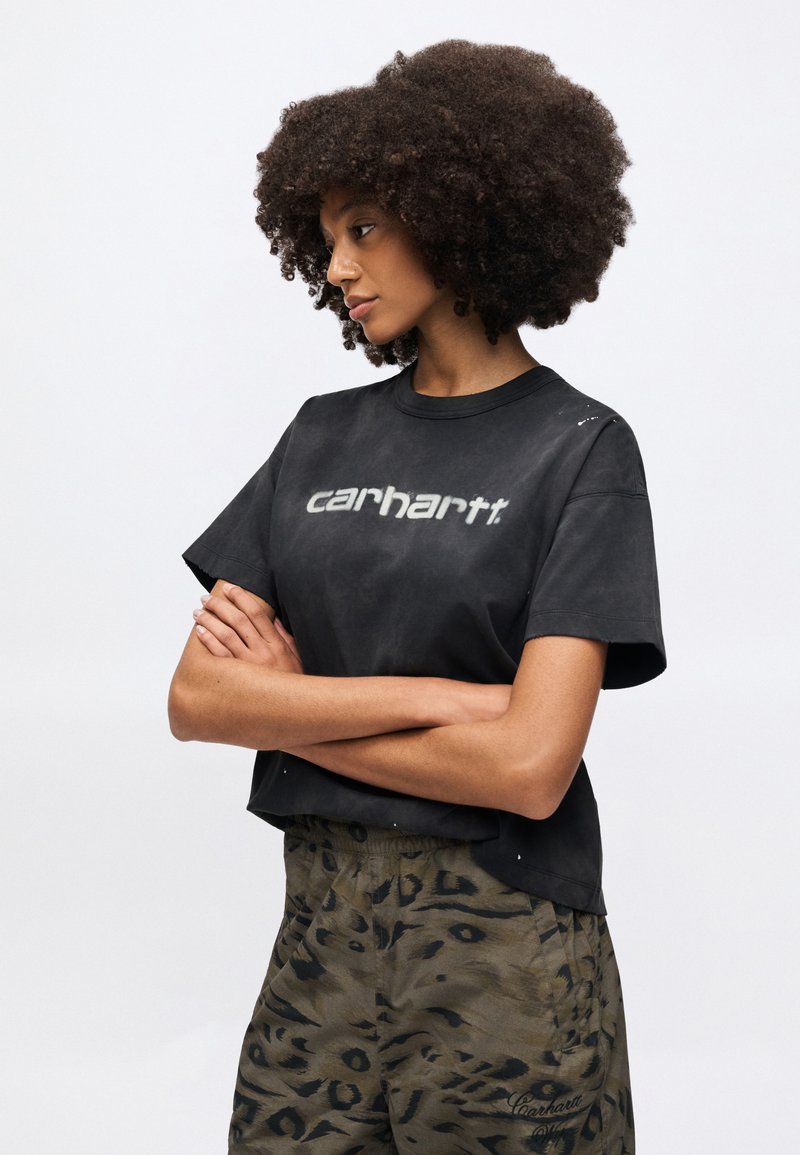 Sort cropped t-shirt med en lysere grå "carhartt" logo, kombineret med grønne bukser med leopardprint, der har en afslappet pasform og korte ærmer.